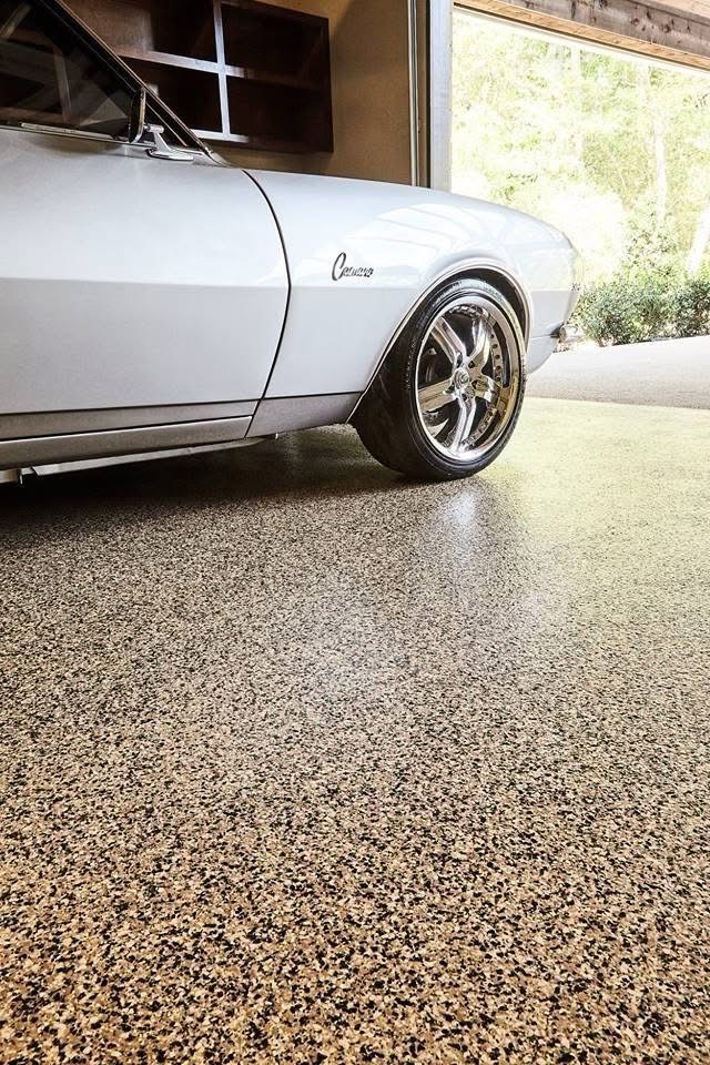 A sleek, silver classic car parked in a garage with a polished, speckled floor reflecting sunlight from an open door.