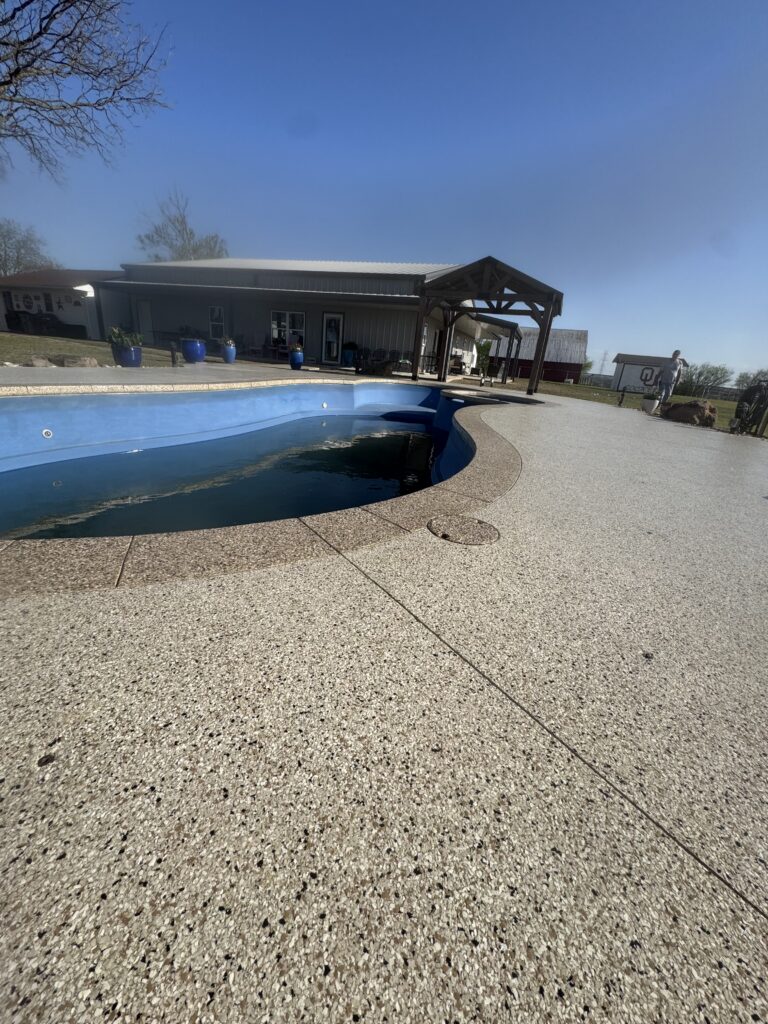 Outdoor swimming pool with a curved edge, surrounded by a speckled concrete deck and a covered pavilion on a sunny day.