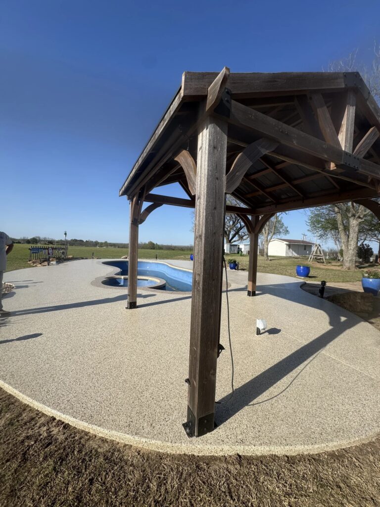 Wooden gazebo in a park with a gravel surface and a small pool in the background under a clear blue sky.
